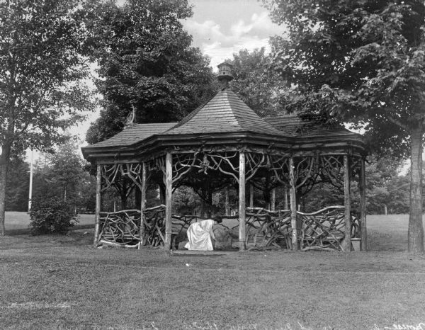 A woman wearing a dress stoops inside a pavilion made of gnarled branches at Deer Park Hotel. Boulders provide seating inside the structure and the shingled roof features a birdhouse at its peak.