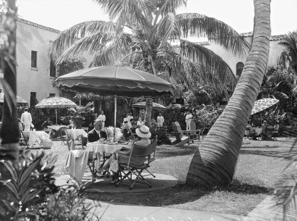 People sit at umbrella-covered tables in the courtyard at the Brazilian Court Hotel. Lush vegetation and palm trees decorate the space and wait staff are in the background.