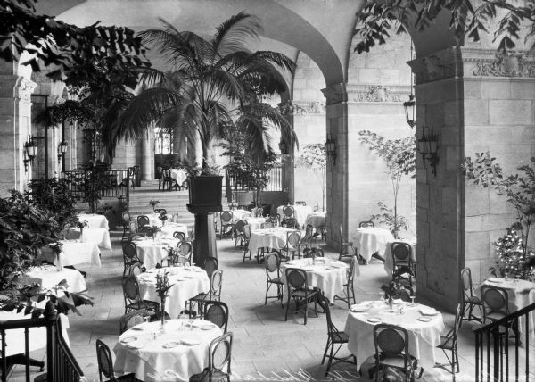 Elevated view of tables in an arcade in the Whitehall Hotel. Each table is covered in cloth, set with plates and glassware, and decorated with a vase of flowers and the arcade is decorated with trees and potted plants.