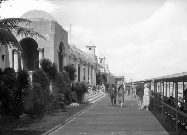 People walk along the Casino Walk promenade between a row of buildings and a beach. Beside the beach is a covered sitting area set with folding chairs.