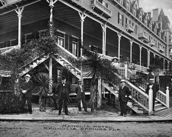 A group of men and women posing in front of the Manolia Hotel for a portrait. The two lowest floors of the hotel are surrounded by porches and the staircase railing leading to the second story is covered in vines. Caption reads: "Manolia Hotel, Manolia Springs, Fla."