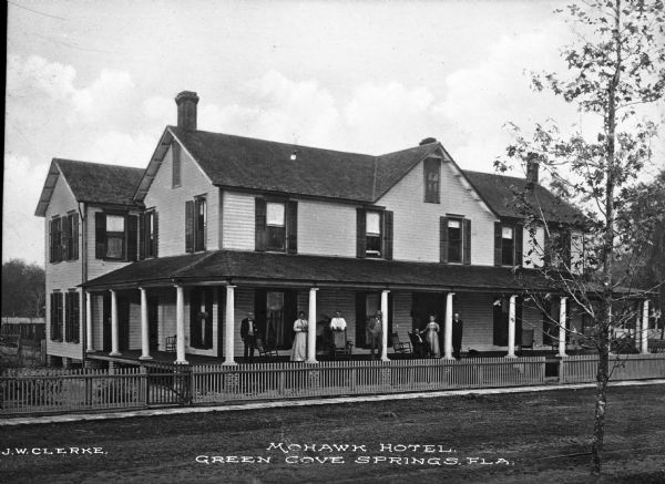 View across road toward a group of men and women posing for a portrait on the porch of the Mohawk Hotel. The building is surrounded by a wooden fence. Caption reads: "Mohawk Hotel, Green Cove Springs, Fla."