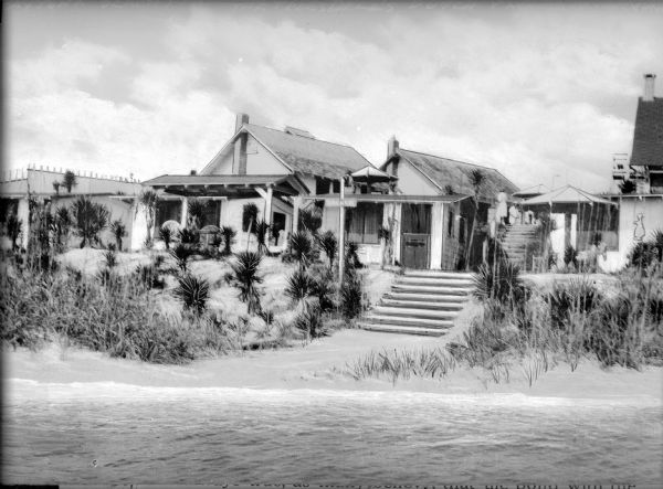 View from water of houses along the beach at Dutch Village, each with small outbuildings and umbrellas set up outdoors. A set of stairs leads from the beach to a courtyard area where men and women have gathered.