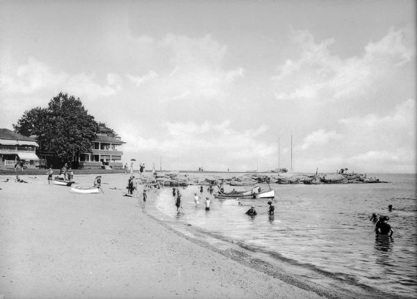 Vacationers wade at a beach near a row of what appear to be hotel buildings. Groups of people sit near boats onshore and a group of women sits at the end of a rock jetty, looking out at the water.