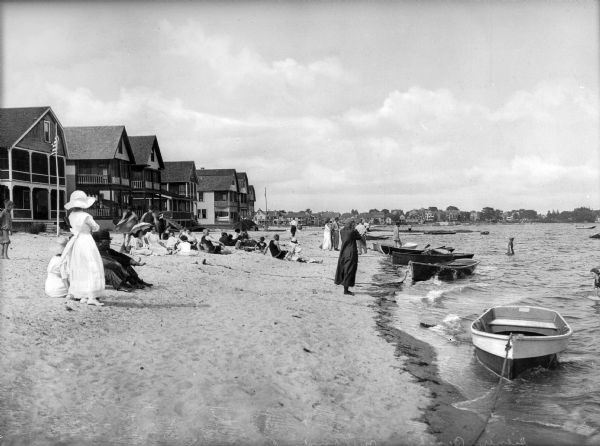 View along shoreline toward people relaxing on a beach lined with vacation houses. Groups of men and women are standing or sitting, some wearing street clothing and some in bathing suits. Other people are wading in the water.
