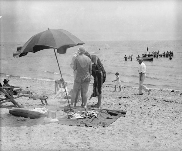A group of sunbathing adults and children standing and sitting near a blanket, folding chairs, and umbrella set up on a beach. In the background, swimmers are gathered near a rowboat and raft out in the water.