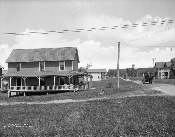 A view across a lawn towards the Palace House, located on Sound View Avenue. Two women and a man sit in rocking chairs on the porch, while children play in the yard. A horse-drawn buggy is parked in the middle of the avenue on the right.