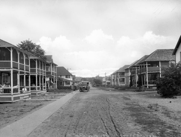 An automobile is driving down a dirt road between two-story houses that line either side of Sound View Avenue. Two children are playing on the sidewalk to the left, and adults are sitting on the buildings' porches.
