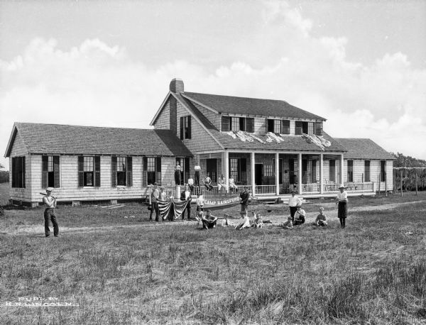 A view of the main lodge at Camp Rainsford. Boys of varying ages stand in front of the structure and pose for the camera. Some hold a Camp Rainsford banner, while others hold a flag. Another group of boys stands on the porch with an adult and blankets and garments appear to be drying on the lodge's rooftop.