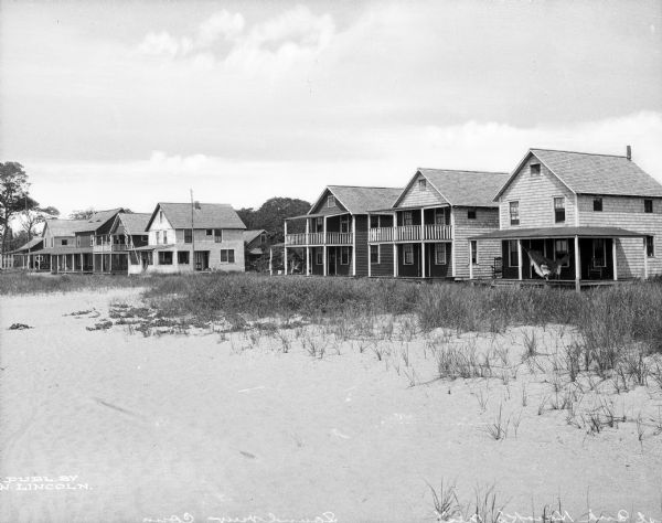 A row of two-story cottages line a beach. To the right, two children are playing in and around a hammock hanging on a porch.