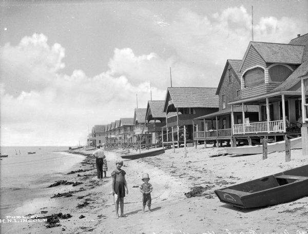 Two children pose for a portrait while standing along a shoreline next to a row of cottages. Several rowboats are pulled ashore, and people are on the cottage porches.