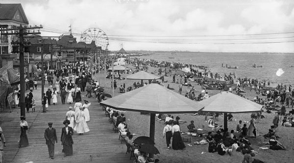 Elevated view of people walking along Midland Beach and boardwalk. Several buildings stand alongside the boardwalk and a Ferris wheel is in the background.