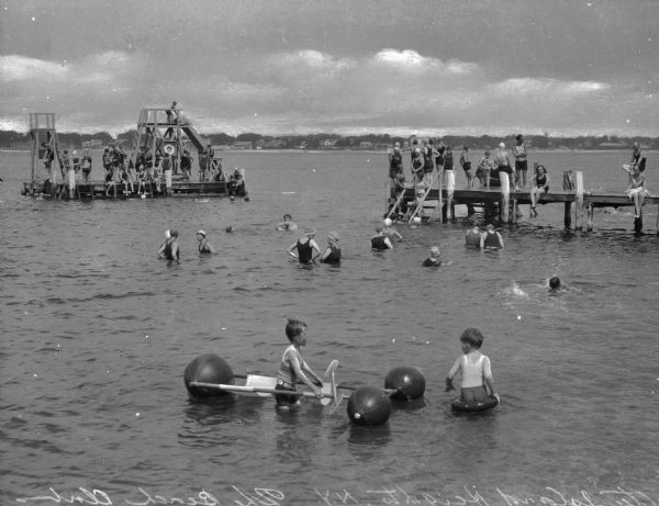 Men, women, and children swim in the water between two docks. Children in the foreground play with a flotation device.
