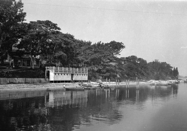 View across water toward the cabins and boathouse at Camp Occiupet. Two boys stand on the shoreline while readying a boat for launch, and another boy sits in the boat behind them, to the right.