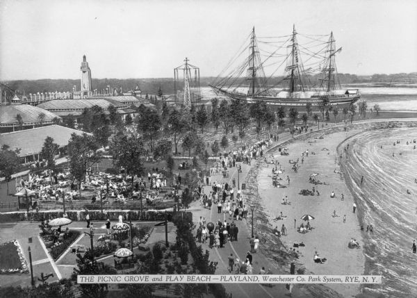Elevated view of the Playland picnic grove and beach, both crowded with people. A large ship, pavilions and outbuildings are in the background. Caption reads: "The Picnic Grove and Play Beach -- Playland, Westchester Co. Park System, Rye, N.Y."