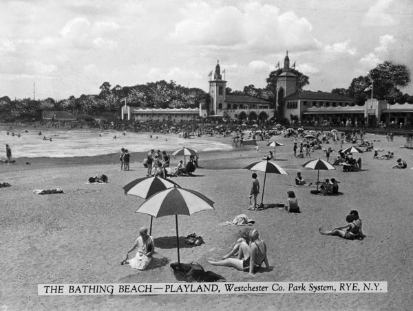 People sunbathe on the beach at Playland while others swim in the surf.  A turreted building decorated with flags is in the background. Caption reads: "The Bathing Beach -- Playland, Westchester Co. Park System, Rye, N.Y."