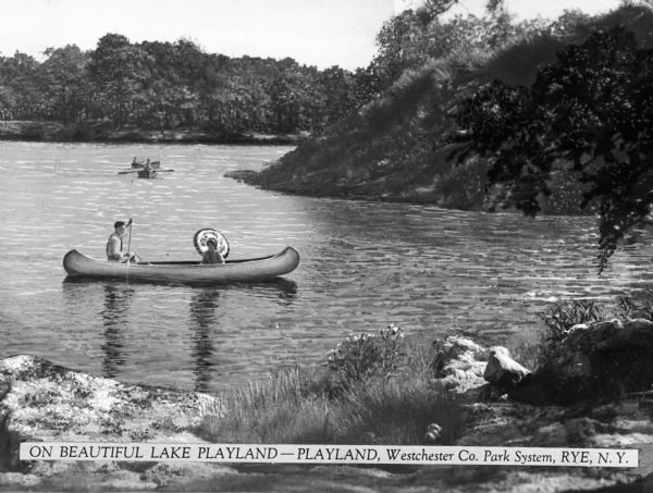 View from shoreline toward a woman in a canoe holding a parasol as an accompanying man paddles through the water near a wooded shore at Playland. Other rowboats are in the background. Caption reads: "On Beautiful Lake Playland -- Playland, Westchester Co. Park System, Rye, N.Y."