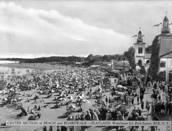 Elevated view of the beach and boardwalk at Playland, crowded with people walking on the boardwalk, sunbathing on the beach and wading in the water. The beach stretches to the horizon, and buildings are along the right. Caption reads: "Center Section of Beach and Boardwalk -- Playland, Westchester Co. Park System, Rye, N.Y."