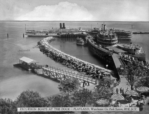 Elevated view from shoreline of excursion boats docked at Playland near a jetty. People walk on the dock to board the boat, and tables shaded by umbrellas are in the foreground at what appears to be a restaurant area. Caption reads: "Excursion boats at the Dock -- Playland, Westchester Co. Park System, Rye, N.Y."