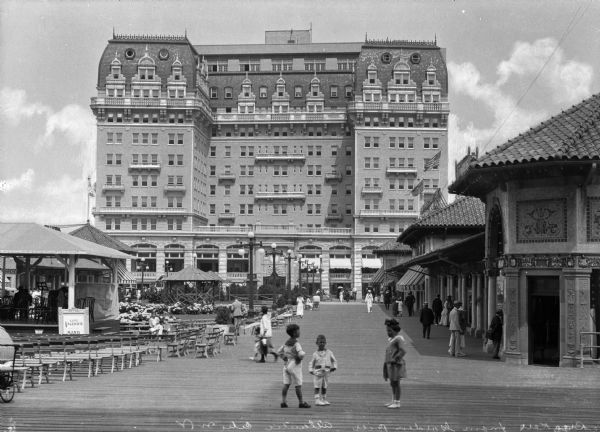 View of the Breakers Hotel taken from the Garden Pier. A pavilion and rows of wooden benches are to the left and a sign advertises "Luigi Valeno's Band." Three children wearing sailor suits are standing in the foreground while others walk along the boardwalk toward the hotel behind them.