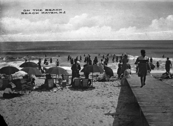 View toward people relaxing on a beach under umbrellas and other people wading in the water. On the right a woman is walking down a boardwalk to get to the beach. Caption reads: "On the Beach, Beach Haven, N.J."