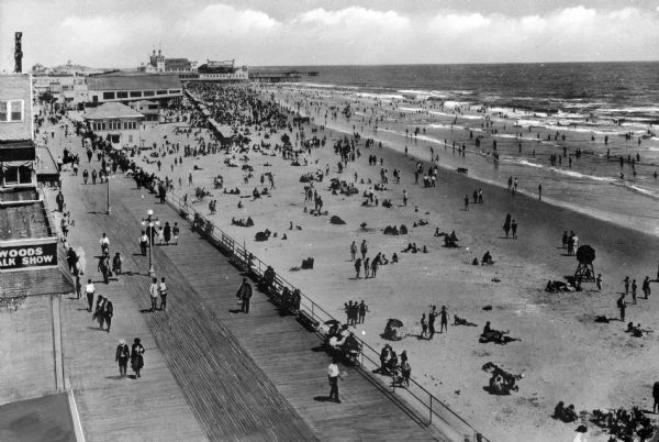 Elevated view of the beach and boardwalk. People are walking down the boardwalk past a line of shops along the left side, and, to the right, sunbathers lie on the beach and others wade in the water.