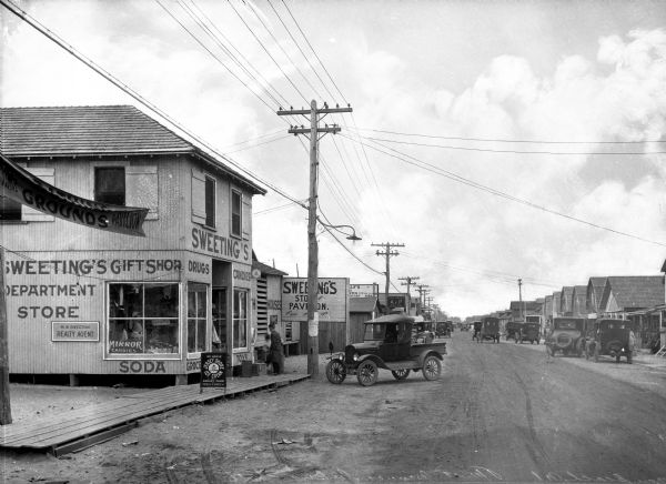 View looking down First Avenue, with shops on the left and small houses on the right. Automobiles are parked along both sides of the street. The signs painted on the building in the left foreground read: "Sweeting's Gift Shop. Department Store. M.W. Sweeting, Realty Agent. Soda. Grocery. Toys. Drugs. Candies." The sign standing on the boardwalk in front of the store reads: "We Serve Jersey Shore Ice Cream. Made in Asbury Park by Grenelle & Schanck, Inc."