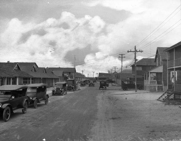 View looking down First Avenue, lined on either side with one and two-story homes. A man stands on the porch of the house in the right foreground and automobiles are parked on both sides of the dirt road.