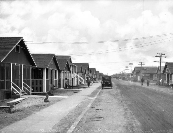 A row of mostly one-story homes standing along First Avenue. A young child plays in the front yard of a home at left while two men walk down the dirt road in the opposite direction.