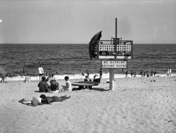 An elevated lifeguard station stands on the sand at Wriests Beach while people sunbathe on the beach and wade in the water. The sign on the lifeguard station reads: "No Bathing Beyond Rope."