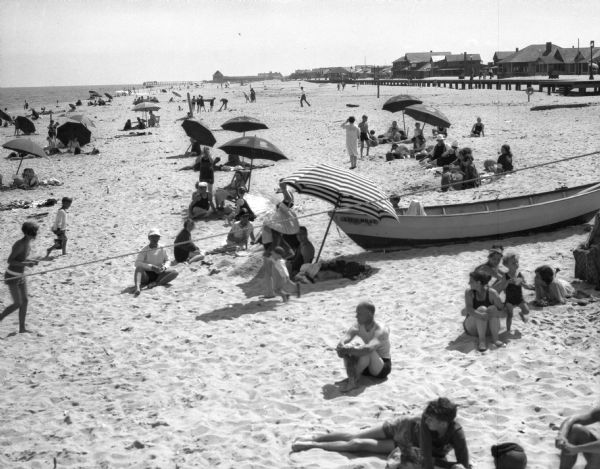 A rowboat rests on the sand at a beach while sunbathers gather around  umbrellas set up nearby. A boardwalk and several buildings are in in the background.