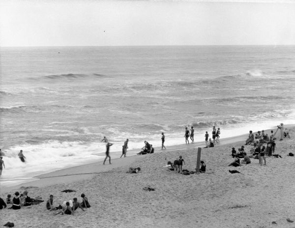 Elevated view toward men, women and children relaxing and sunbathing on the beach. A few people are wading in the water.