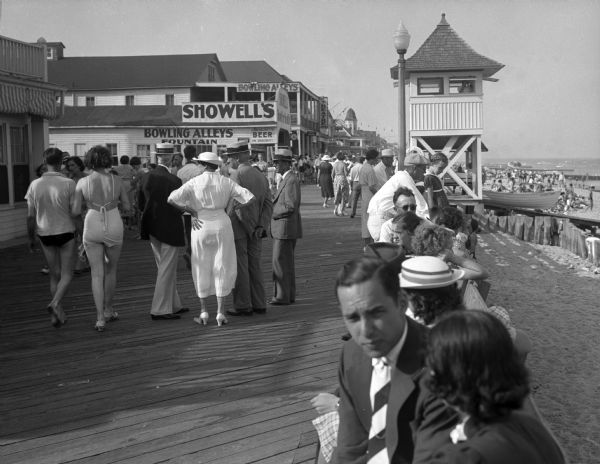 View along boardwalk, with people sitting, and other people walking.  Buildings are along the left, and on the right is a lifeguard tower that looks down onto the beach along the right. The sign on the building to the left advertises "Showell's Bowling Alleys. Fountain, Beer."