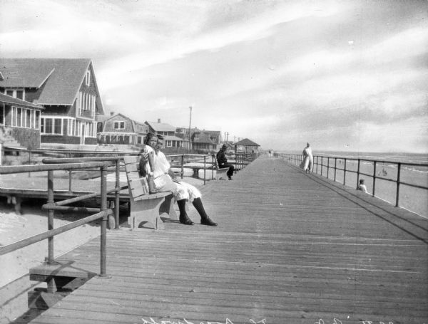 View down wide boardwalk, with a man and a young girl sitting on a boardwalk bench on the left. Several other people look out at the ocean from the railing, and other people are sitting on benches in the background. Houses are along the left.
