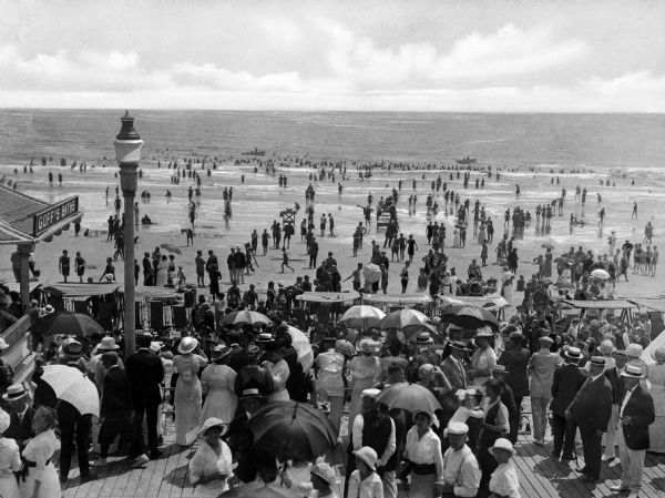 Elevated view of a large crowd of people gathered on the boardwalk. In the background people are walking on the beach, and walking and swimming in the ocean.