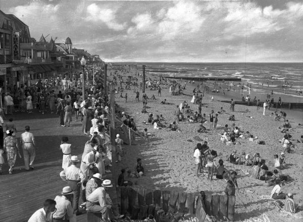 People crowd the boardwalk at Tenth Street, entering and exiting the shops at left. On the right people are sunbathing on the beach and swimming in the surf.