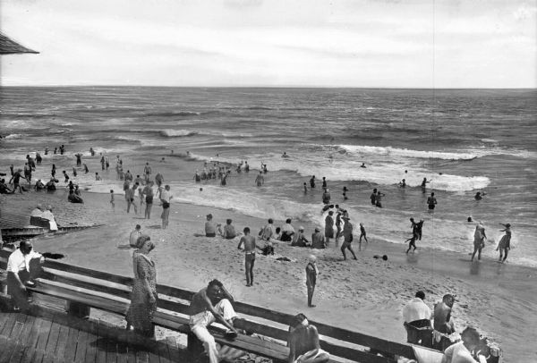 Elevated view of people relaxing on benches in the foreground, and people sitting on the beach and wading in the water along the seashore.