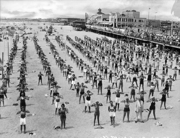Elevated view of men, women and children standing in long rows along the beach, with fingers touching their shoulders, caught in the middle of an exercise movement. Along the right a crowd on the boardwalk observes them.