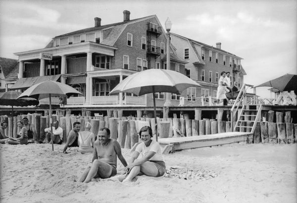 Men and women sit beneath umbrellas on the beach in front of the Colonial Hotel. In the background people are walking on an elevated boardwalk.