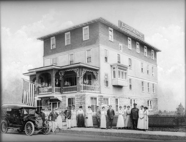 Men and women stand on the sidewalk in front of the Loraine Hotel near a parked automobile. One of the men holds a pennant reading, "Ocean City Teachers Welcome." Another man and woman stand on the first floor porch decorated with small American flags and striped shades; a larger flag projects outward from the porch.
