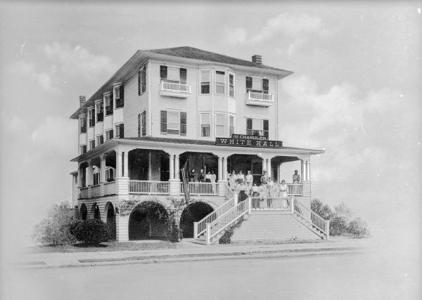 Front view of the Chandler White Hall Hotel. A double staircase leads to the first story porch where a group of people, mostly women, pose.