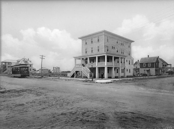 View toward the Shore Hotel from across the intersection at 34th Street and Central Avenue. A double staircase leads to the second-story porch where three children are standong. A trolley is on the dirt road to the left.