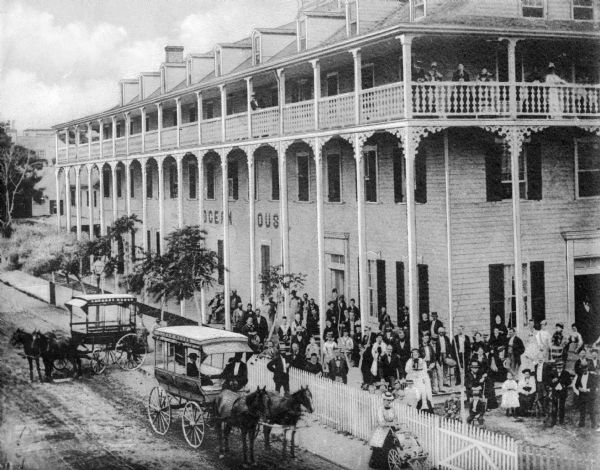 Elevated view of the Ocean House hotel with men, women, and children posing in front of it. Another group of people are standing on the third story balcony. Two horse-drawn carriages are in the dirt road along the front of the hotel.