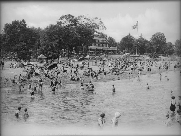 Elevated view across water towards couples, adults, and children sunbathing on the beach and wading in the water. In the distance is an establishment with a name written on its awning, appearing to read: "The Bay House."