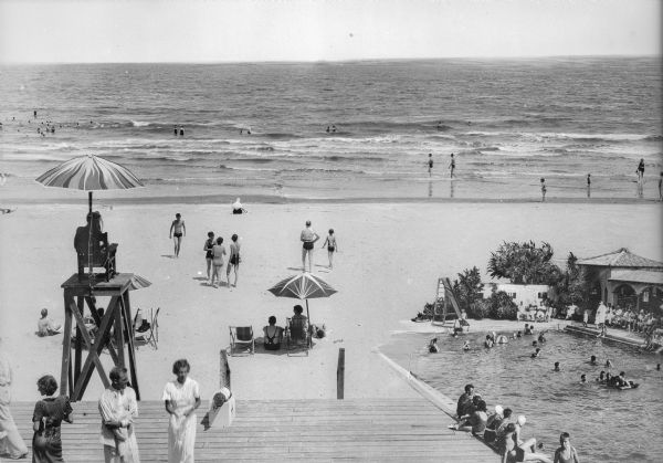 Individuals swim and sunbathe on a beach while others use a swimming pool located beside it. In the foreground, a lifeguard sits on an elevated platform near the edge of a wooden platform.