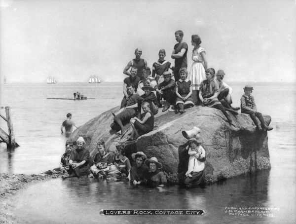 Adults and children posing on and in front of Lover's Rock, which is partially submerged in water. Several sailboats are on the horizon. Caption reads: "Lovers' Rock, Cottage City."