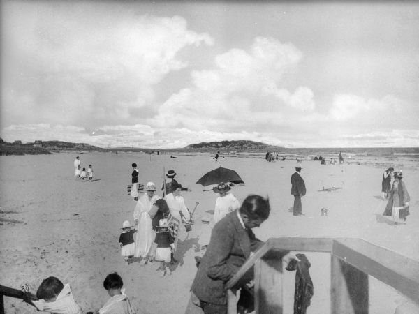A view of the shore and ocean at Good Harbor Beach. People in the foreground are using a set of wooden steps to exit the beach area while other people are walking on the beach or are sitting beneath umbrellas in the background.