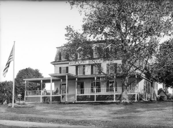 Front view of the Oceanic Hotel as seen from the street. Three people sit on the front porch of the hotel and an American flag flies from a pole in the front yard.