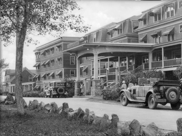 View from lawn toward two automobiles parked in front of the Lookout Hotel. A covered entrance over the staircase leads to the building's porch. Several people are in front of the building.