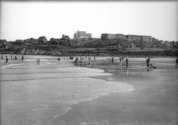 View along seashore toward people playing on the beach and wading into the water during low tide. Buildings line the hills in the background.